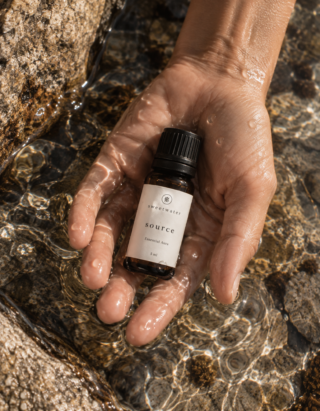 Source Essential Aura 5ml amber bottle held in a hand beneath clear mountain water