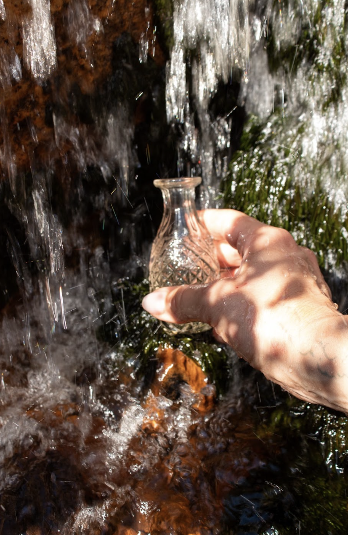 A hand holding a small glass vessel beneath a waterfall, gathering water from the stream