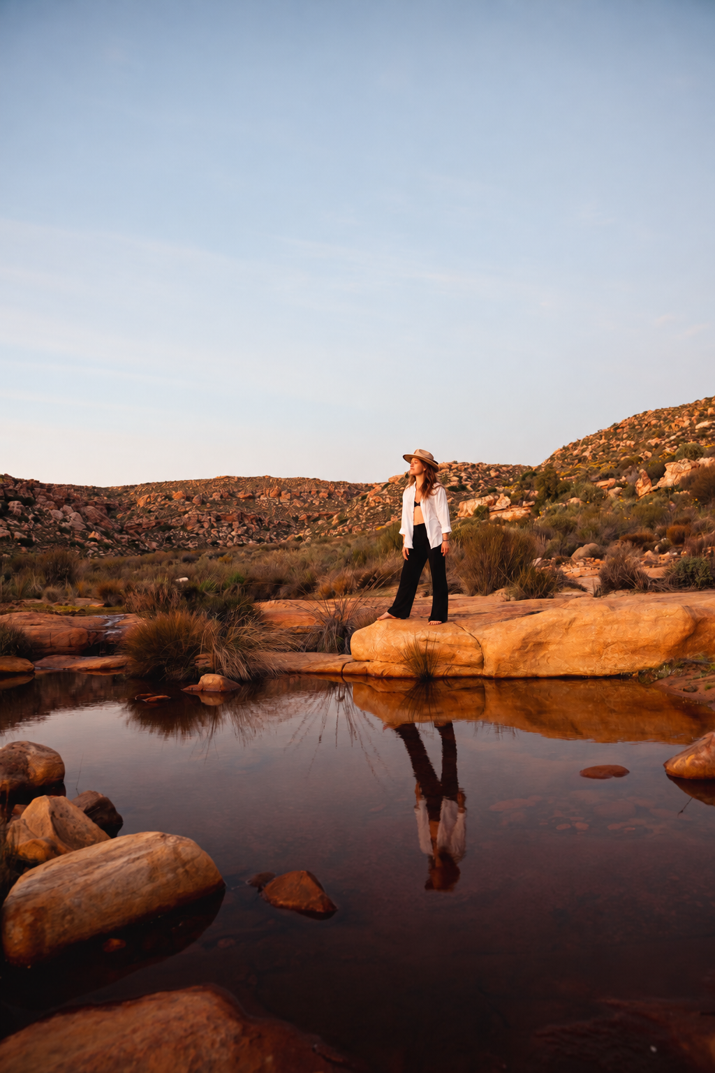 Mikaela standing barefoot on sunlit rocks at the edge of a still mountain pool at dusk, her reflection resting on the water